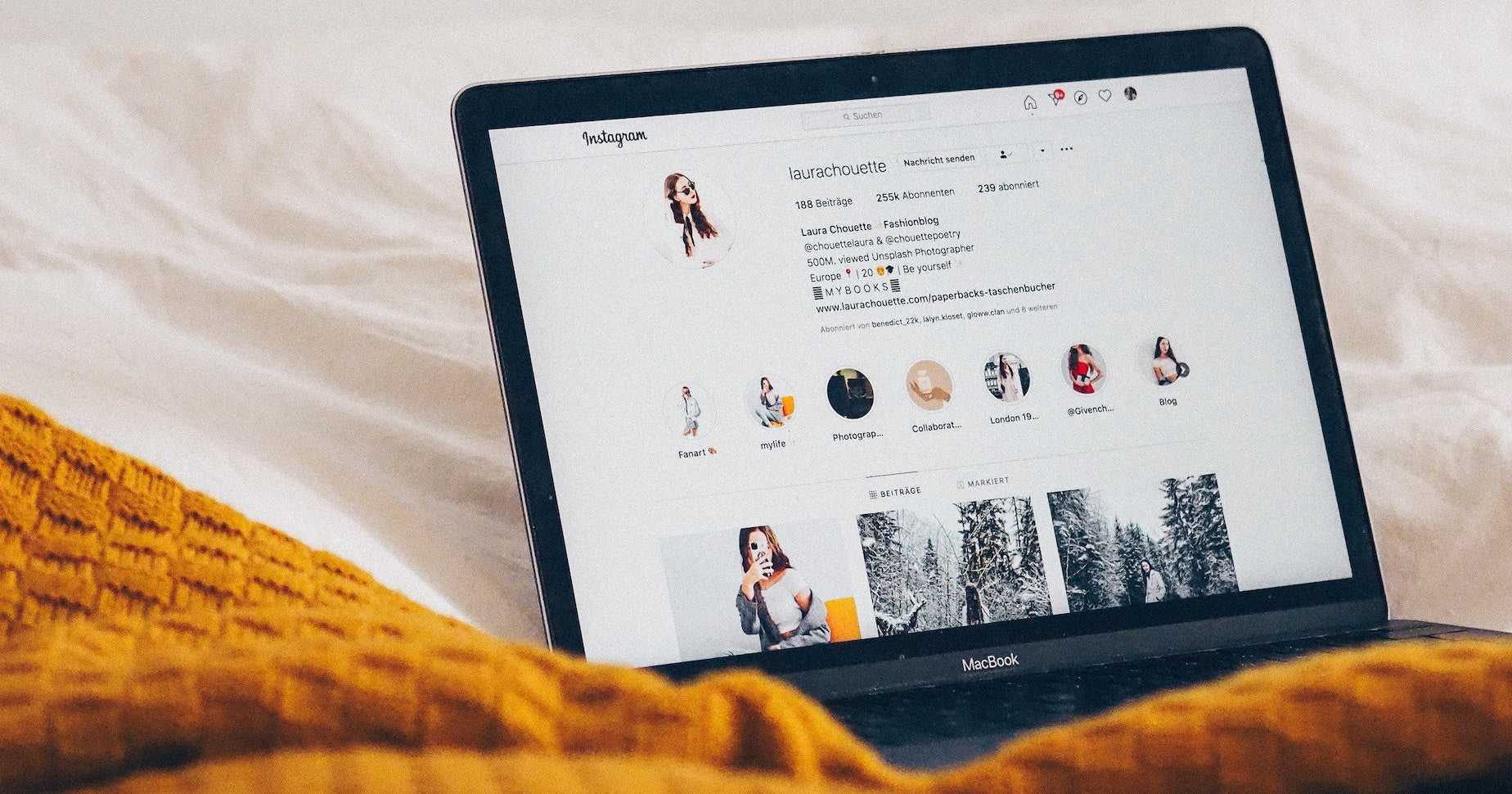A laptop sits open to an influencer's Instagram page on a cream and rust bedspread